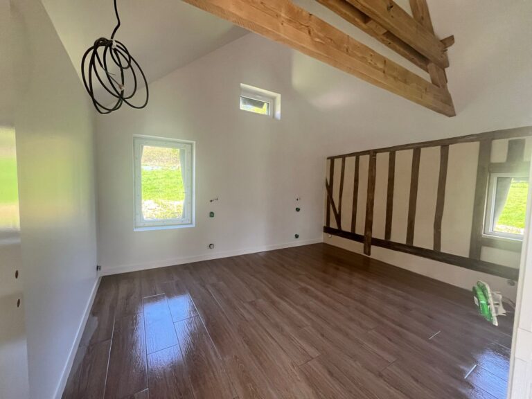 Pièce rénovée avec colombages normands apparents en mur de fond, poutres chêne en hauteur, parquet stratifié foncé et murs blancs, rénovation maison à pans de bois