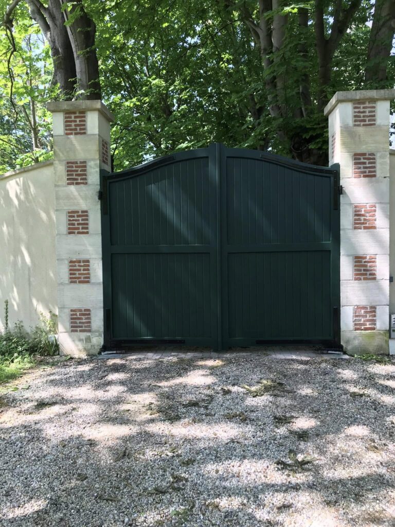 **Balise alt :** ``` Portail double battant en bois vert foncé entre deux piliers en pierre et brique, allée en gravier et arbres feuillus en arrière-plan ``` --- **Variantes :** **Courte :** ``` Portail en bois vert foncé sur piliers pierre et brique, entrée de propriété avec allée gravillonnée ``` **SEO (si contexte menuiserie/rénovation extérieure) :** ``` Portail en bois peint vert bouteille avec piliers en pierre de taille et brique, réalisation sur mesure en Normandie