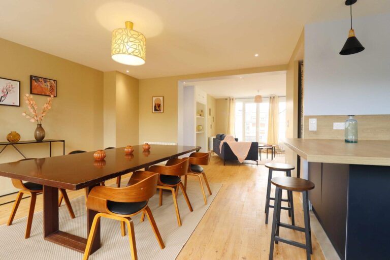 Salle à manger à Lisieux en Normandie, ouverte sur salon avec grande table en bois foncé, chaises scandinaves en bois courbé, parquet clair et cuisine ouverte avec îlot, tons ocre et bleu marine