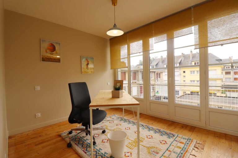 Bureau lumineux avec parquet en bois clair, grande fenêtre à battants donnant sur des immeubles, chaise ergonomique noire, table en bois blanc et tapis berbère coloré. Peinture & Décoration Intérieure à Lisieux.