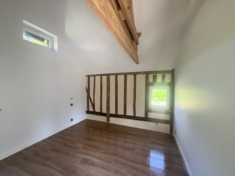 Pièce rénovée avec colombages normands apparents en mur de fond, poutres chêne en hauteur, parquet stratifié foncé et murs blancs, rénovation maison à pans de bois