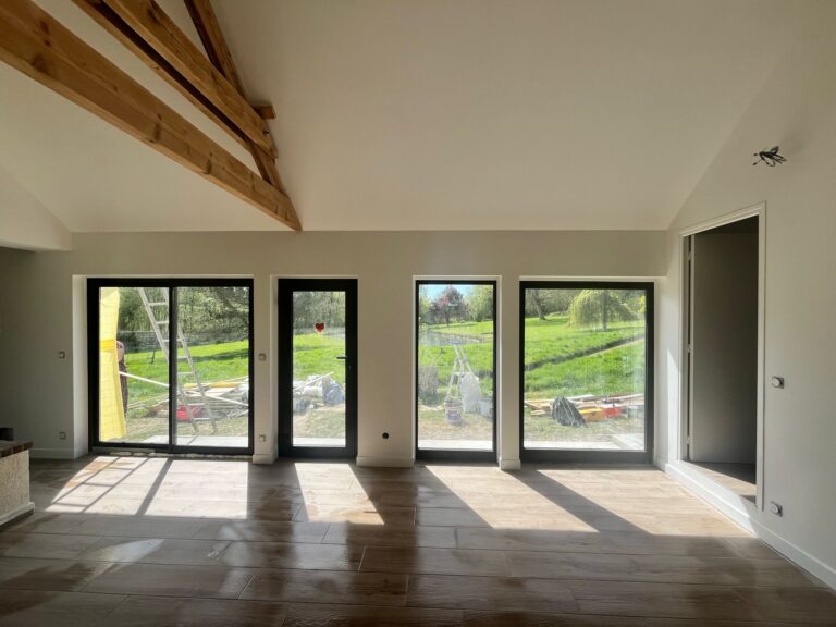 ￼￼Grande pièce de vie en chantier avec baies vitrées et portes-fenêtres noires sur jardin verdoyant, poutres chêne apparentes en hauteur et carrelage imitation bois, construction neuve en Normandie