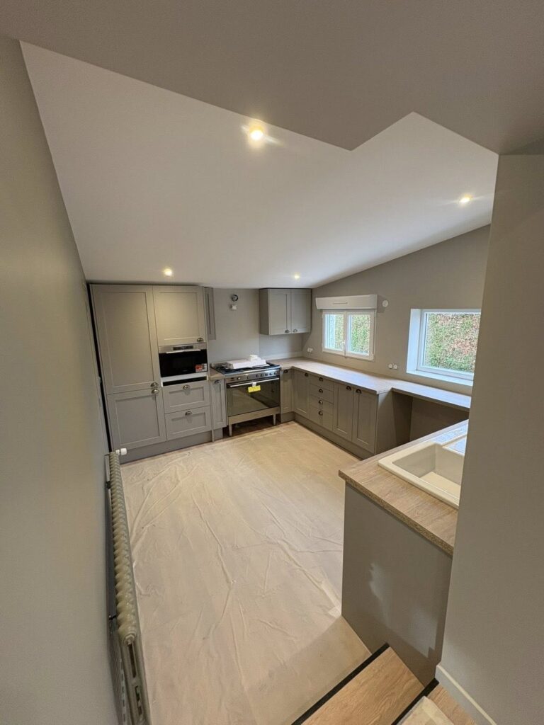 **Balise alt :** ``` Cuisine en L en cours d'installation avec meubles gris taupe style shaker, piano de cuisson inox, spots encastrés et plafond en pente, chantier en fin de travaux ``` --- **Variantes :** **Courte :** ``` Cuisine shaker gris taupe en L avec piano de cuisson, fin de chantier rénovation ``` **SEO (agencement/rénovation) :** ``` Installation cuisine sur mesure style shaker en Normandie, meubles gris taupe en L avec piano de cuisson inox et spots encastrés